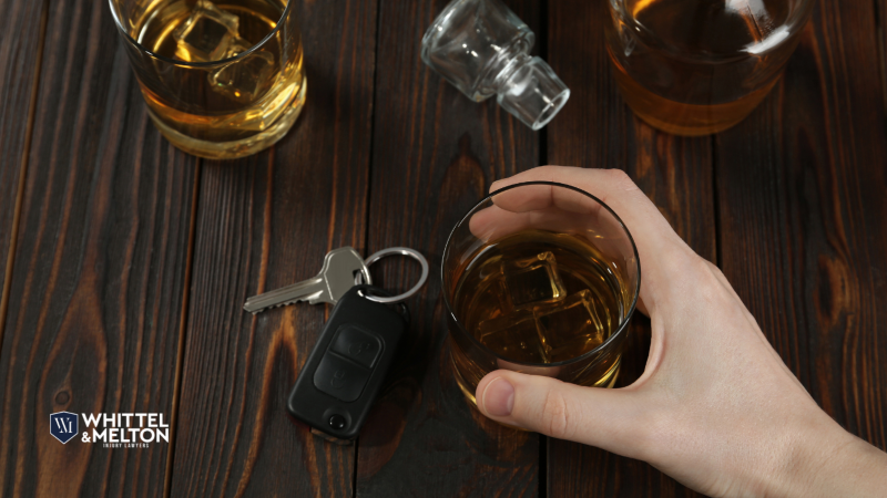 Hand holding a glass of whiskey next to car keys on a wooden table, symbolizing the dangers of drinking and driving.