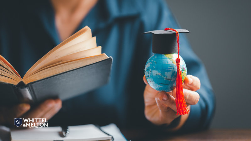 A person holding an open book in one hand and a small globe with a graduation cap in the other, symbolizing education and global opportunities, with Whittel & Melton's logo in the bottom left corner.